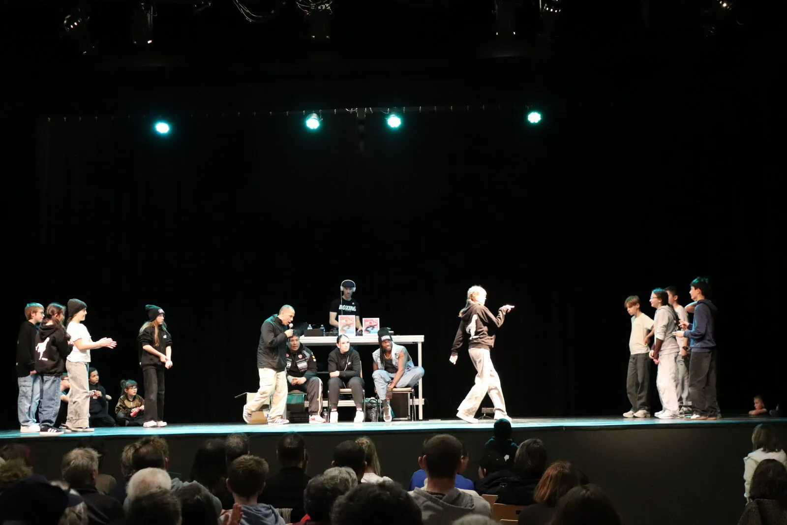 Battle de breaking sur scène : danseurs en confrontation, animateurs et DJ, public en premier plan